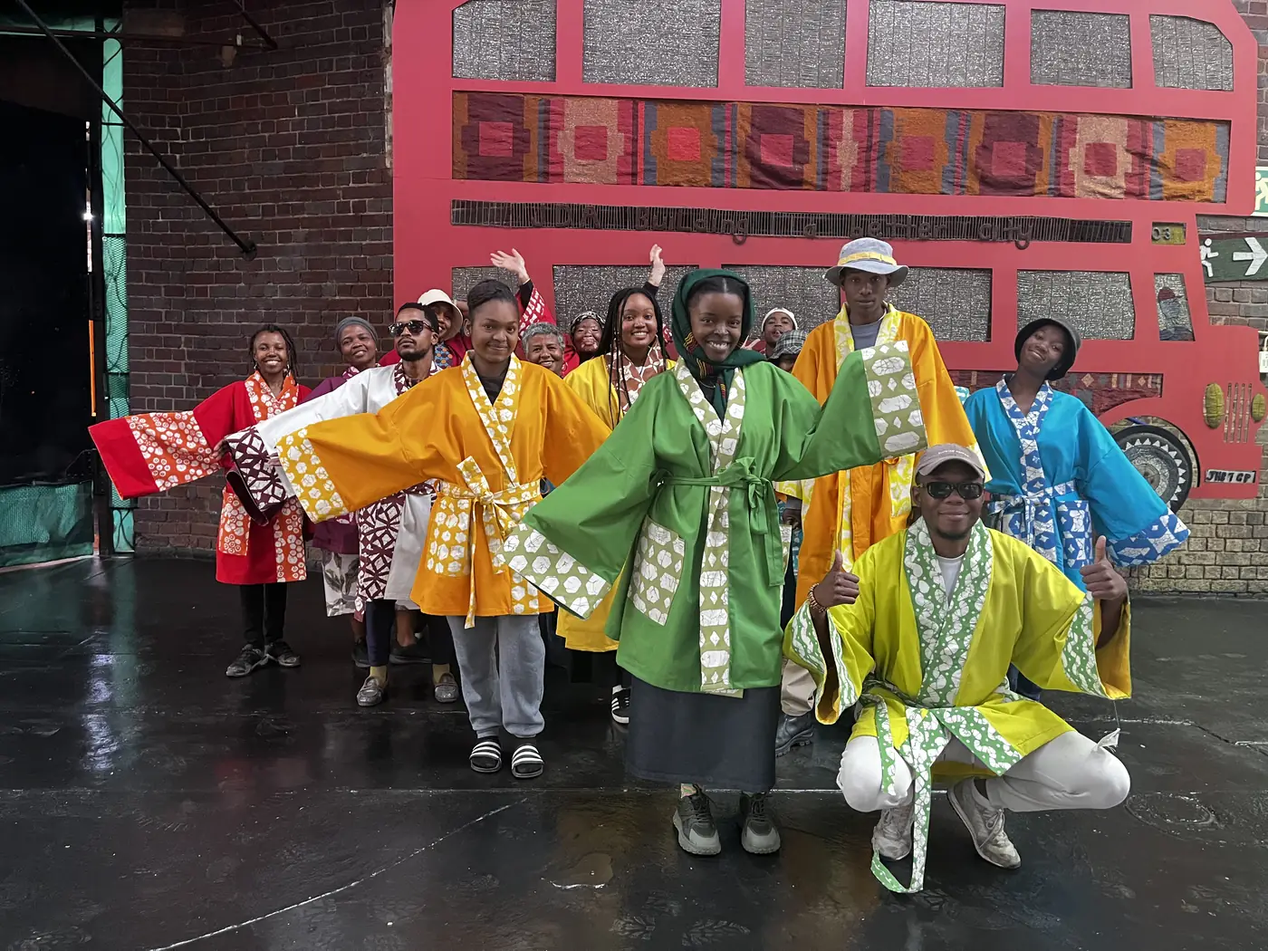 Imbali students in bright orange, green, yellow and blue jackets made from their own printed fabric, smiling and waving in front of a red bus mural
