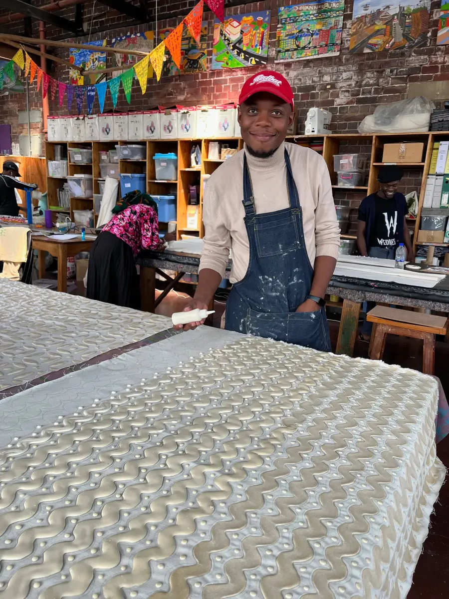 A maker in a red cap and denim apron smiles beside a long print table of freshly screen-printed cloth
